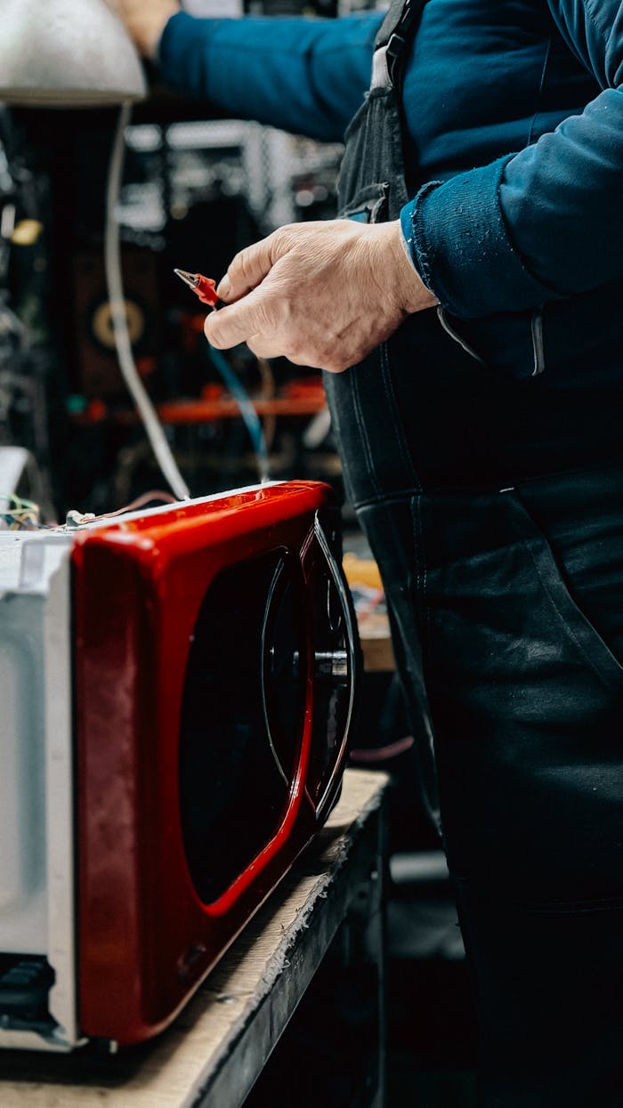 Electrician working on red microwave repair in a workshop setting, tools in hand.